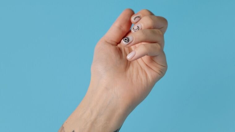 Close-up of a tattooed hand with artistic nail art against a pastel blue background, showcasing creativity and style.