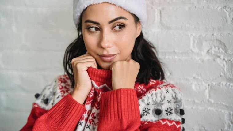 Young dreamy ethnic female with brown eyes wearing warm winter sweater and Santa hat and looking away