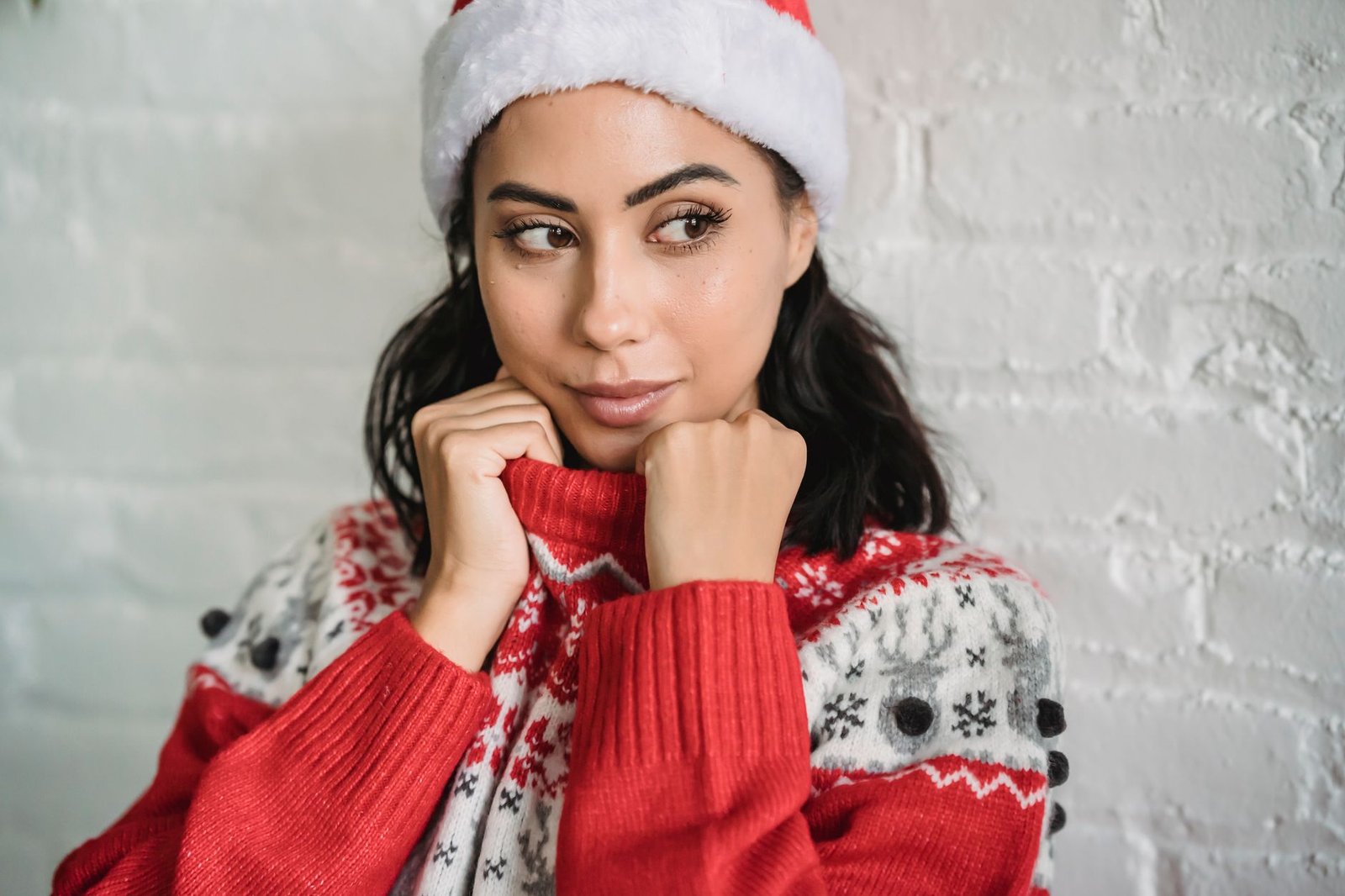 Young dreamy ethnic female with brown eyes wearing warm winter sweater and Santa hat and looking away